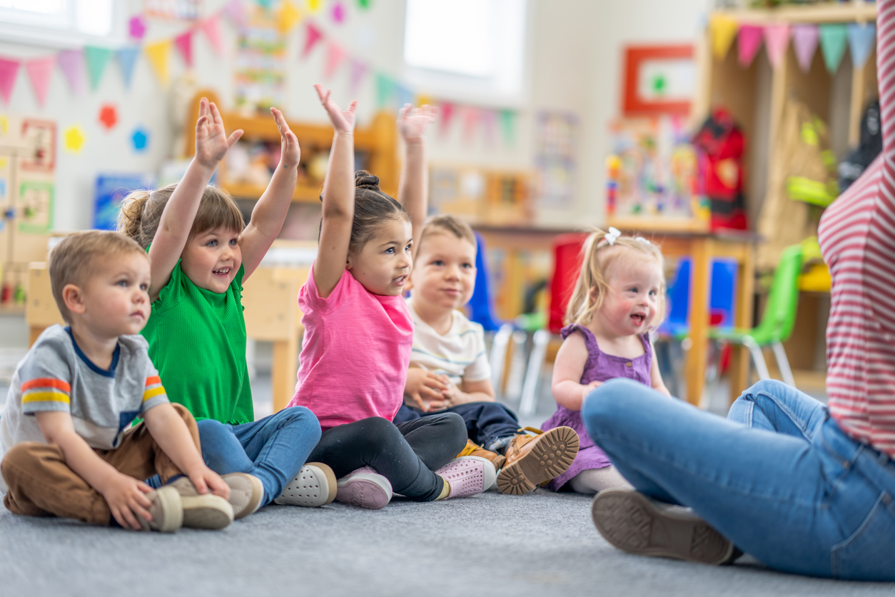 A preschool teacher sits on the floor of her classroom with her students in front of her as she leads them in a sing-along. The children are dressed casually and smiling as they following along with the actions.